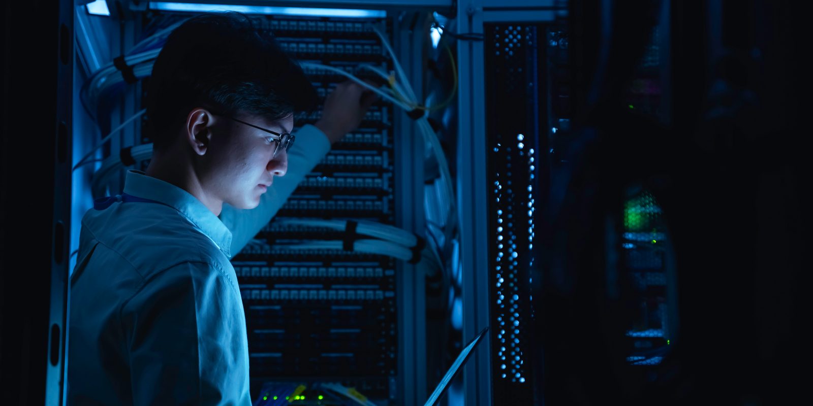 A cyber engineer inspecting server hardware with a tablet in a dark data center representing hands-on cyber engineering and infrastructure management