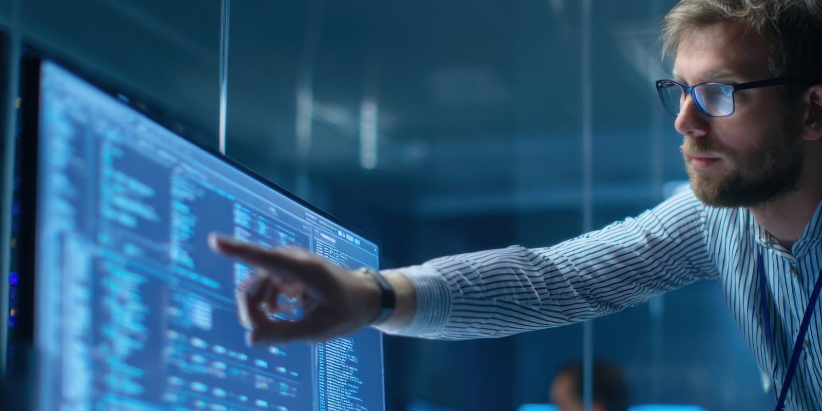 A cybersecurity professional pointing at a large monitor displaying system data in an operations center representing active cybersecurity management and program