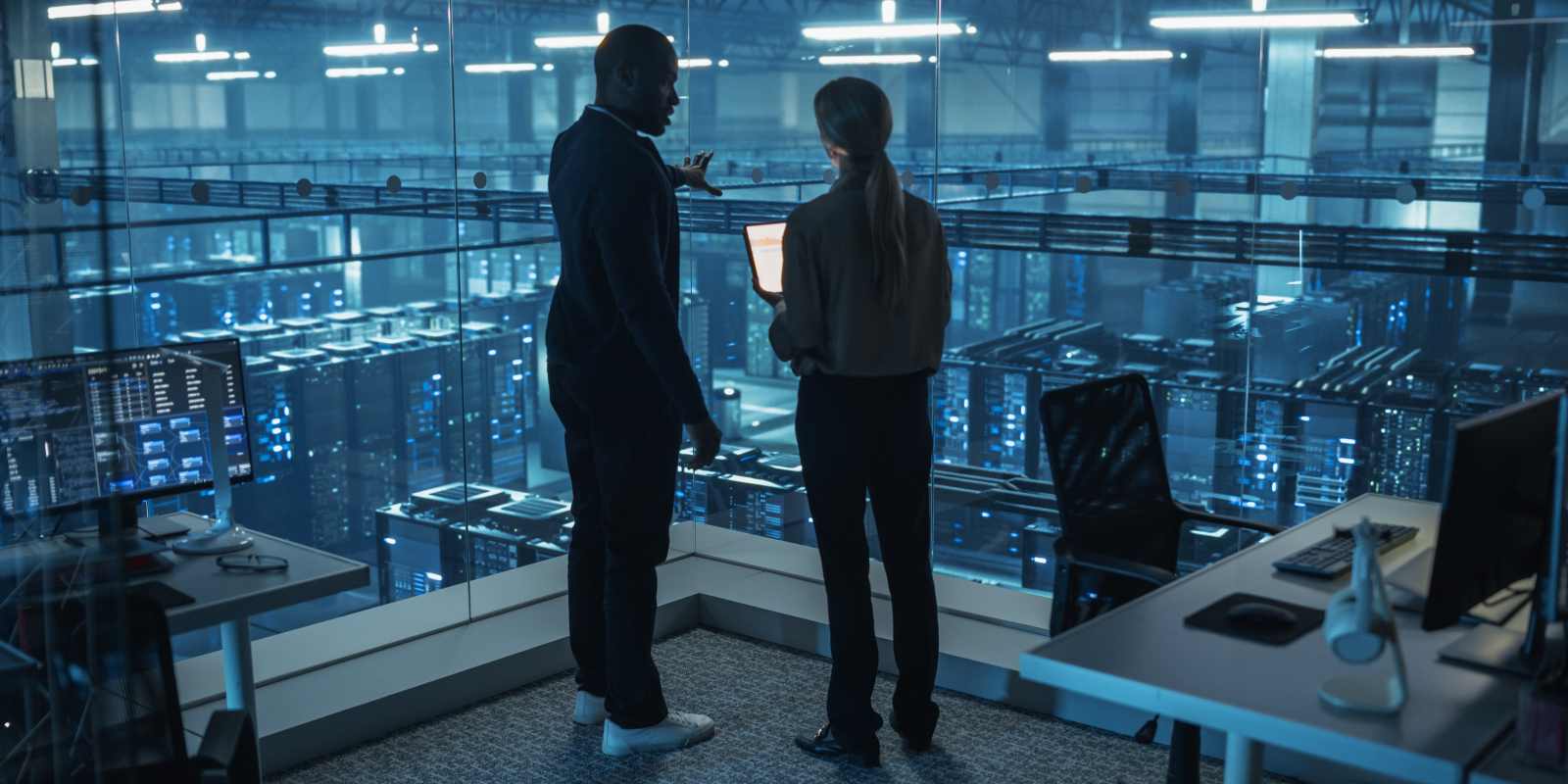 Two cybersecurity consultants discussing over a laptop overlooking a large data center representing professional cybersecurity consulting
