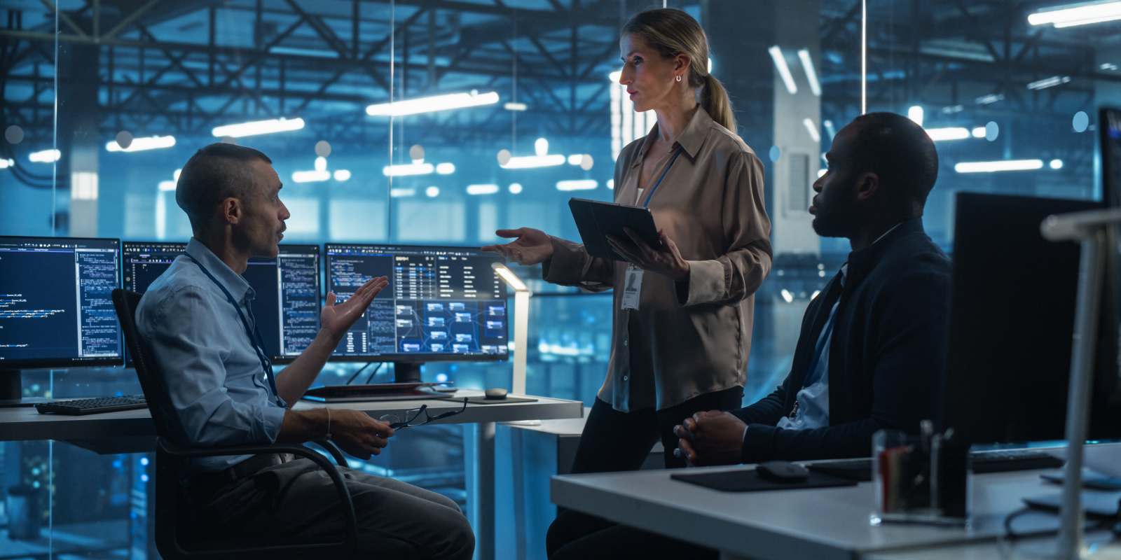 diverse cybersecurity team discussing findings around workstations with code on multiple monitors representing collaborative cybersecurity operations