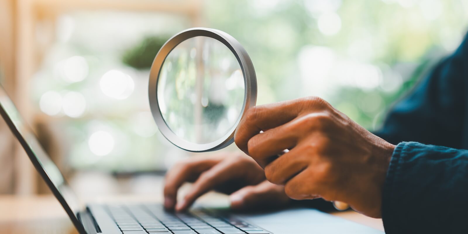 person using magnifying glass while working on laptop, representing cybersecurity analysis