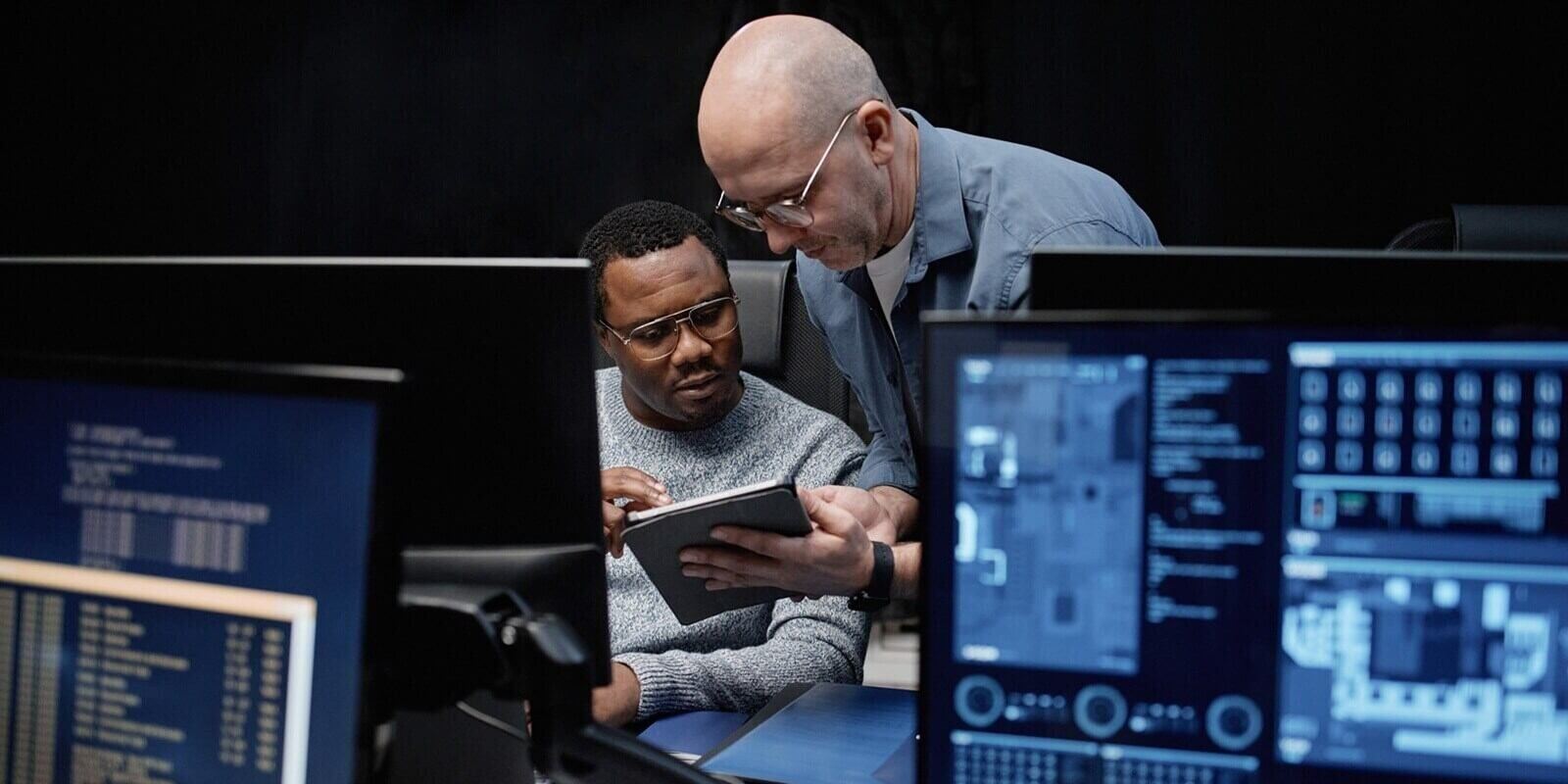 caucasian man showing digital tablet to young Black man while sitting at workstation with multiple computer monitors